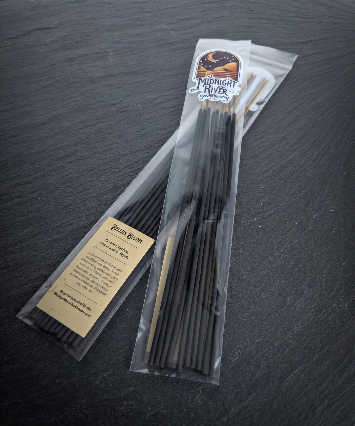 Clear bag of black hand dipped incense sticks with a Midnight River Apothecary Bellus Besom label, lying on a dark slate surface.