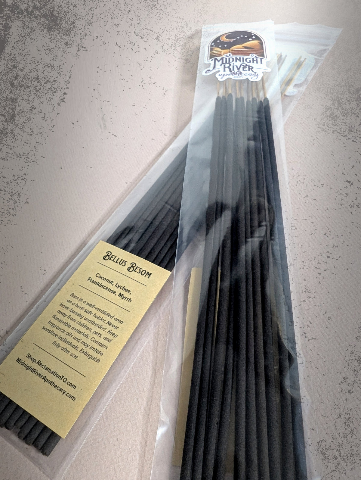 Second angle of the Bellus Besom incense bag from Midnight River Apothecary on a dark surface, with the yellow label and the black sticks inside.