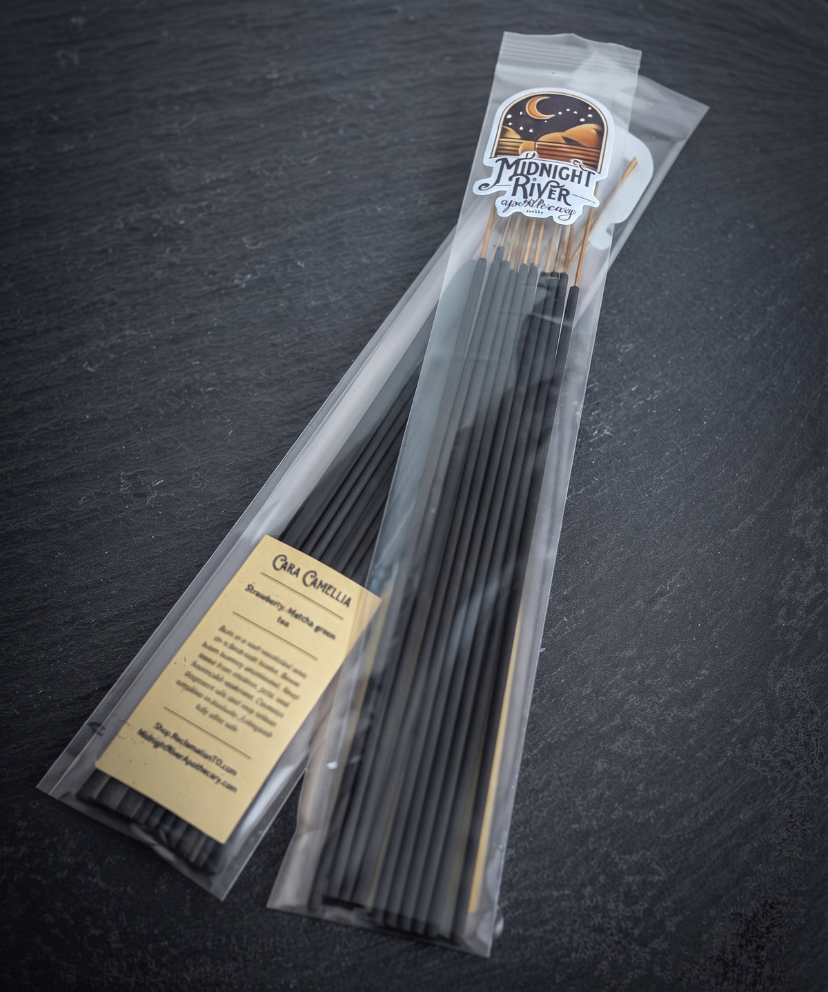 Pack of Cara Camellia hand dipped incense by Midnight River Apothecary lying at an angle on dark slate, with the cream label reading “Cara Camellia” on the clear bag and black sticks inside