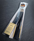 Angled view of the bagged black incense sticks on dark slate, with the cream ingredient label at the lower left