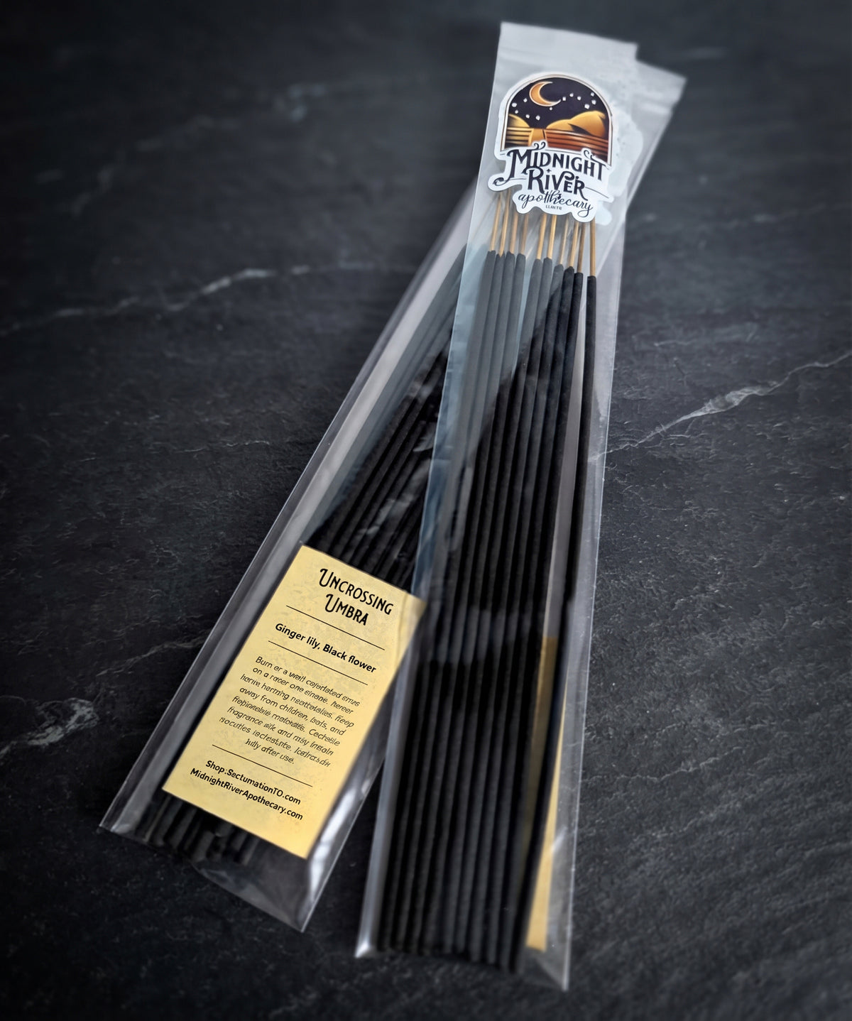 Clear bag of black hand dipped incense sticks with a Midnight River Apothecary Uncrossing Umbra label, lying on a dark slate surface.