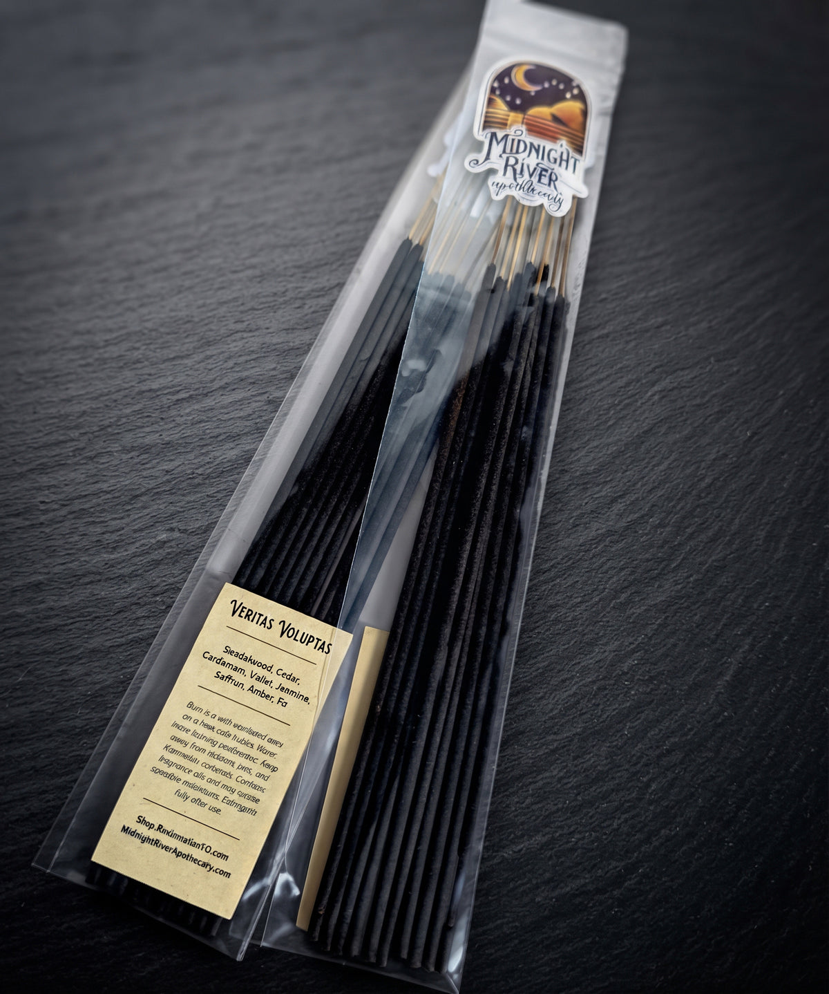 Pack of Veritas Voluptas hand dipped incense by Midnight River Apothecary lying at an angle on dark slate, with the cream label reading “Veritas Voluptas” on the clear bag and black sticks inside