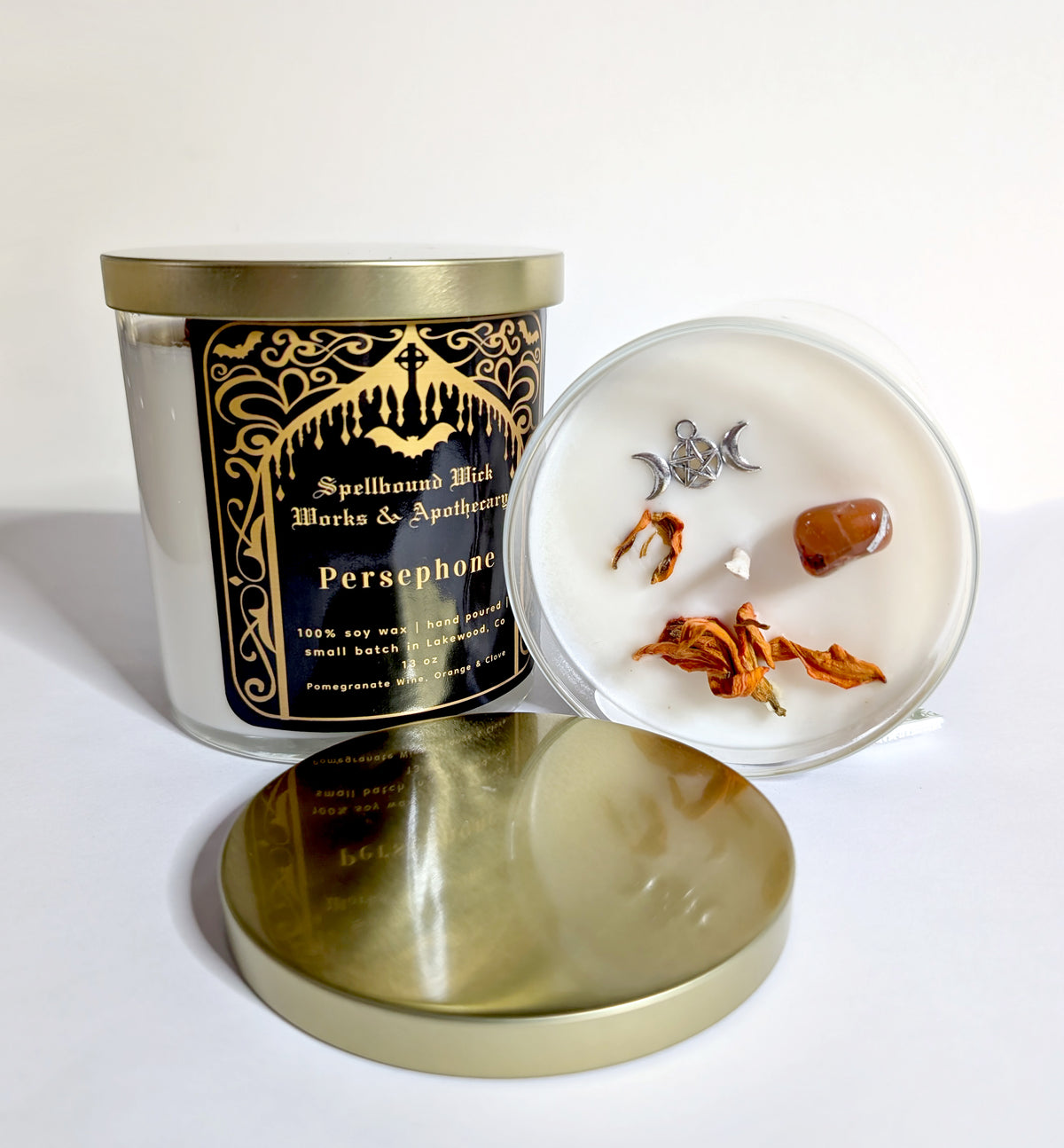 The Persephone Goddess Candle with its lid propped beside it, showing the triple moon charm and dried botanicals set into the white wax