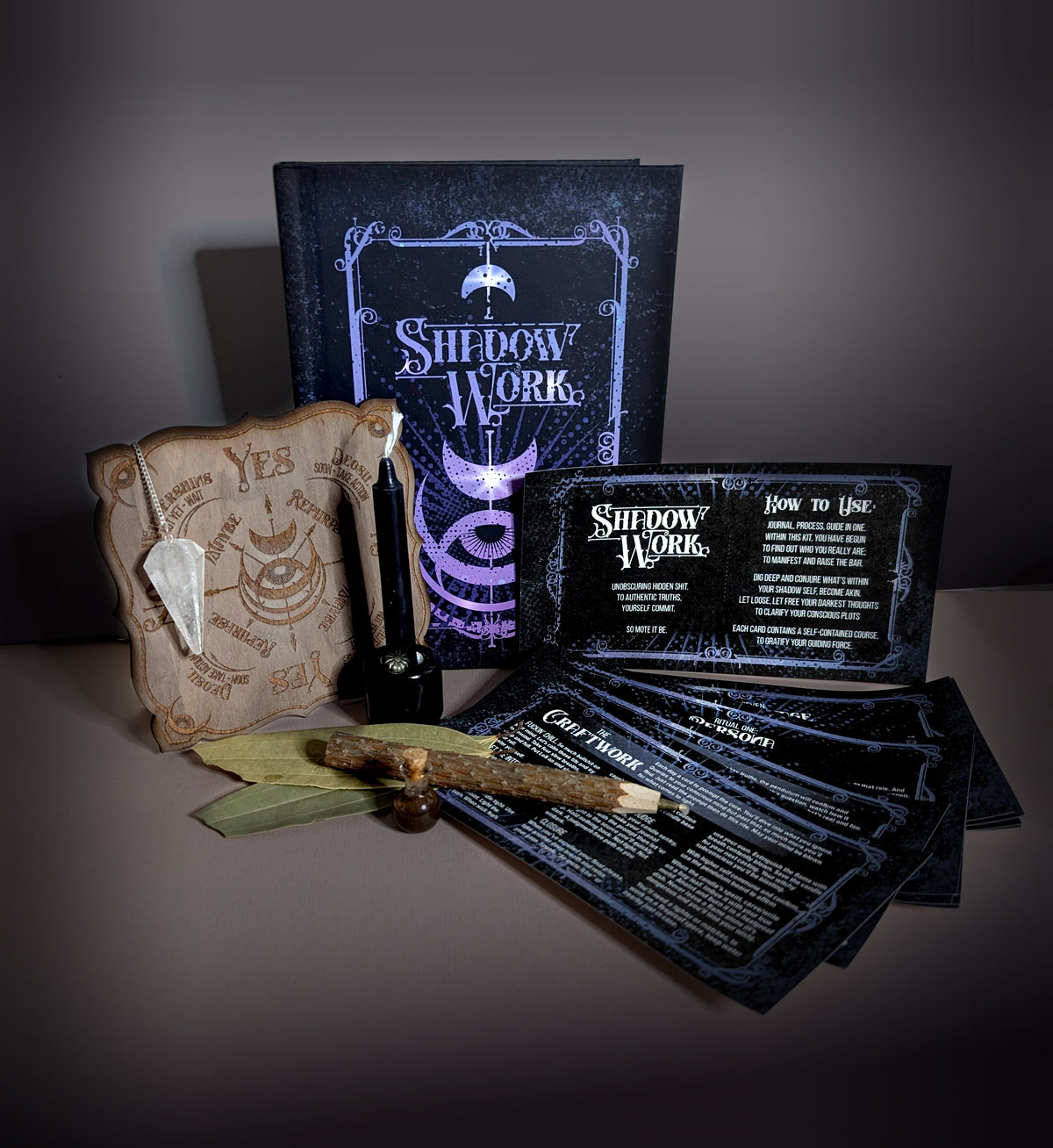 The full Shadow Work Ritual Kit laid out on a pale background, with the journal, pendulum board and quartz pendulum, black chime candle in its holder, bay leaves, twig pencil and the fanned card set