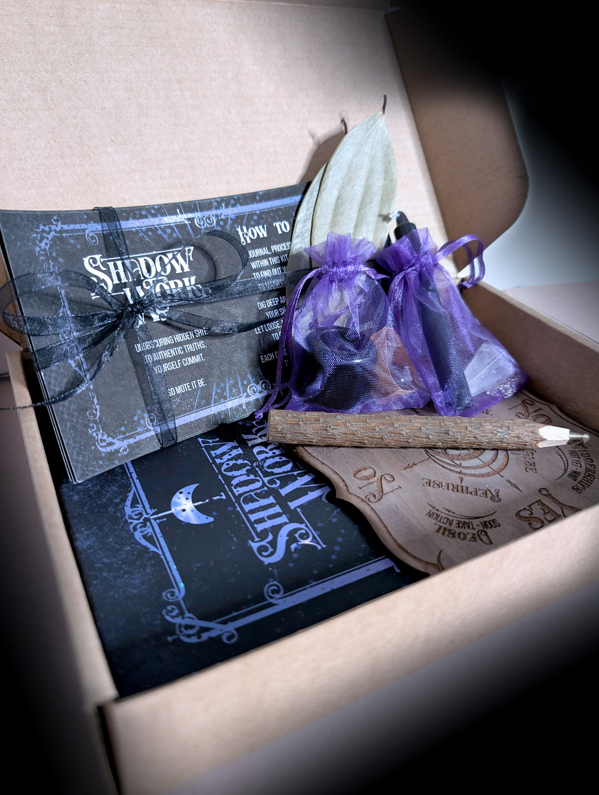 Close view inside the open Shadow Work kit box, showing the ribbon tied cards, the purple organza bags, the twig pencil and the etched pendulum board