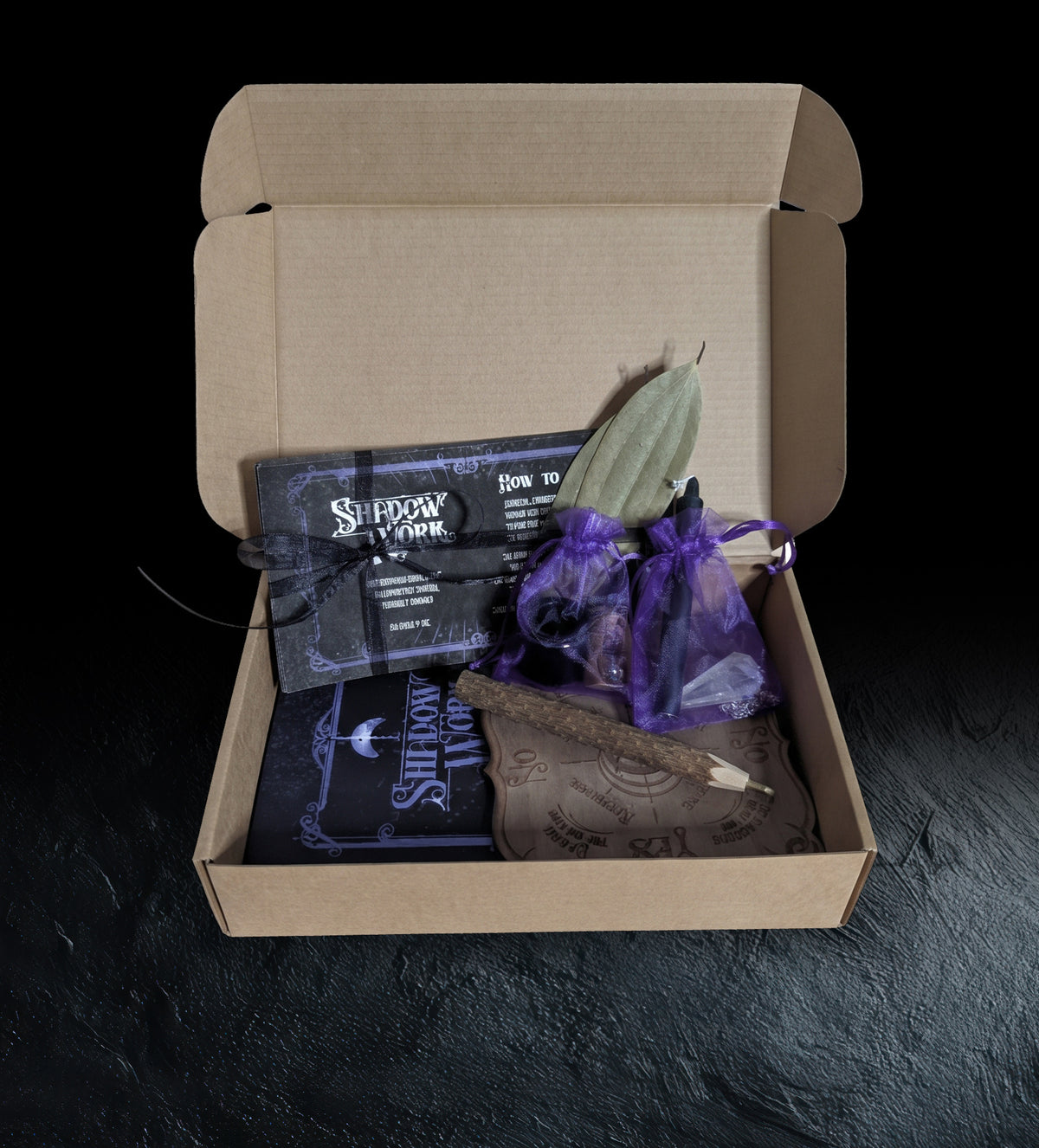 The Shadow Work Ritual Kit packed in its kraft box, with the card set tied in black ribbon, two purple organza bags holding the pendulum and candle, a bay leaf, the twig pencil, the pendulum board and the journal