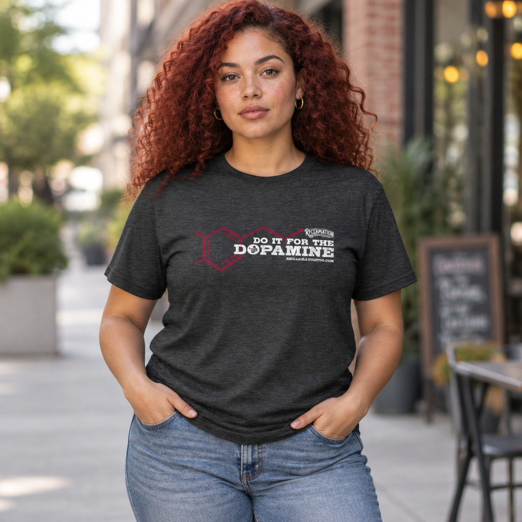 Model wearing the Do It For The Dopamine t-shirt in dark grey heather on a city sidewalk, the dopamine molecule and lettering printed across the chest in dark red and deep purple