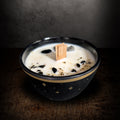 Black bowl candle with gold stars painted on the outside, seen from the front on a dark background, cream soy wax set with black and snowflake obsidian around a wooden wick