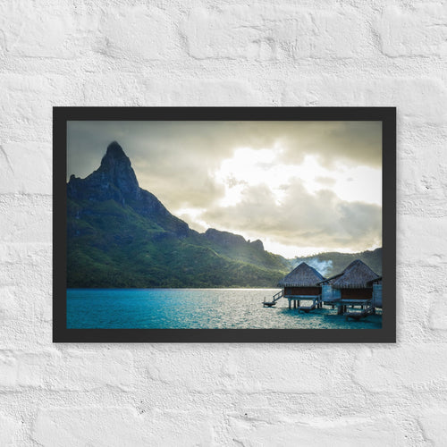 Bora Bora, French Polynesia framed luster print by iain, black framed photograph of Mount Otemanu rising over turquoise water with overwater bungalows under heavy clouds, hanging on a white brick wall