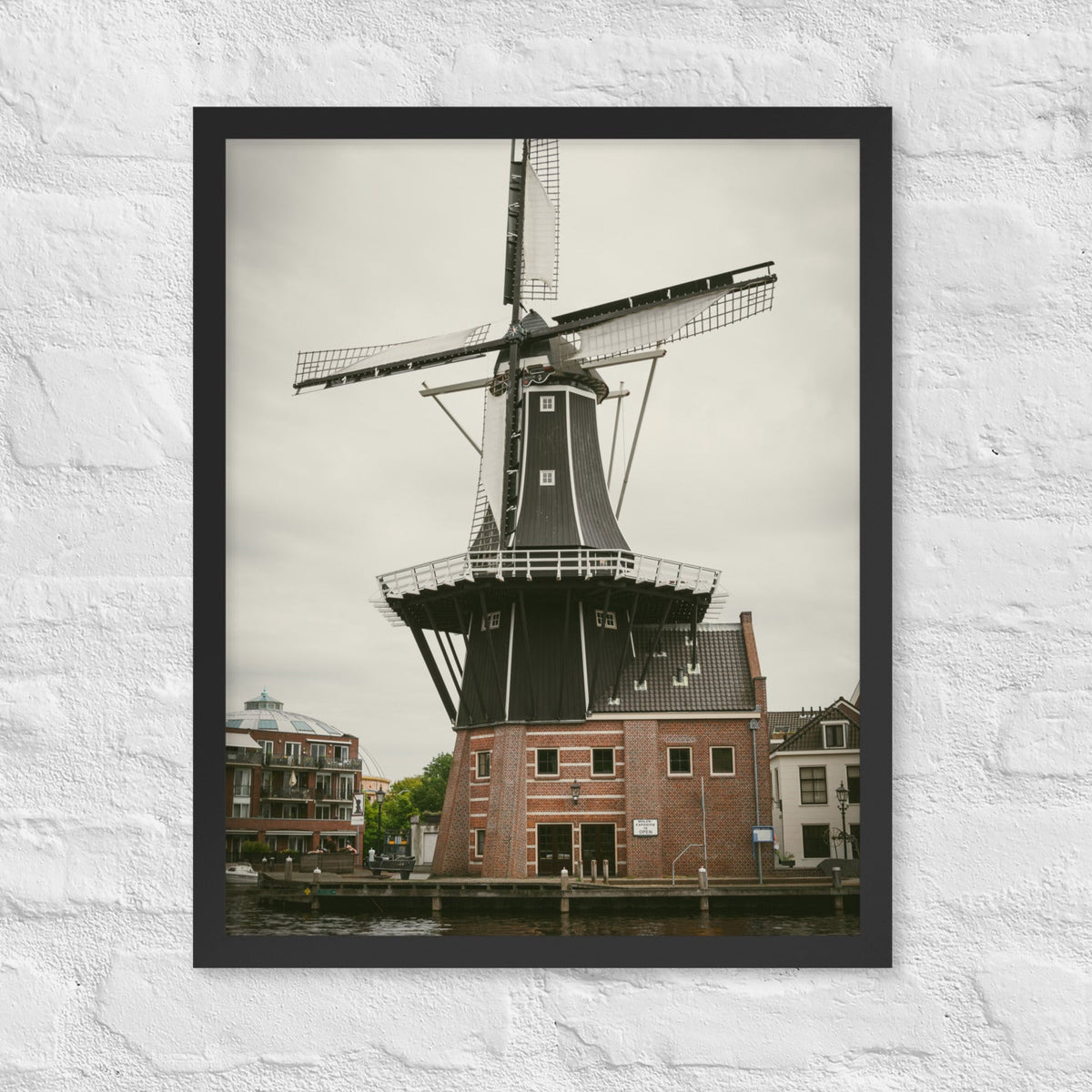 De Adriaan, Haarlem framed luster print by iain, black framed photograph of the De Adriaan windmill rising above brick buildings under an overcast sky, hanging on a white brick wall