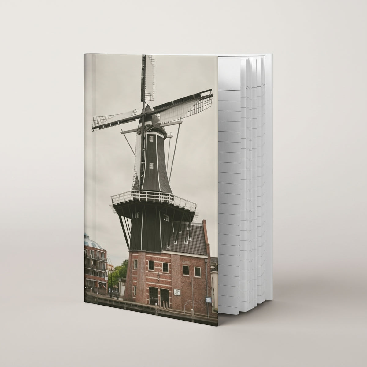 De Adriaan Windmill hardcover journal by iain, journal standing upright on a soft neutral background, with a photograph of the De Adriaan windmill in Haarlem and its brick building on the front cover and the page edges showing on the side