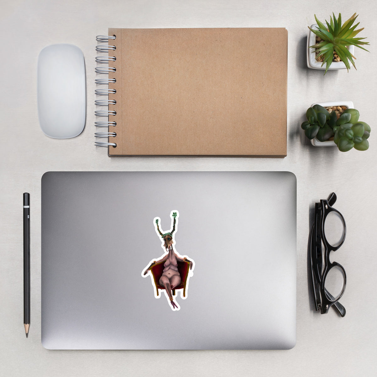 Matte vinyl sticker applied to the lid of a silver laptop on a desk setup with a notebook, pen, and glasses.