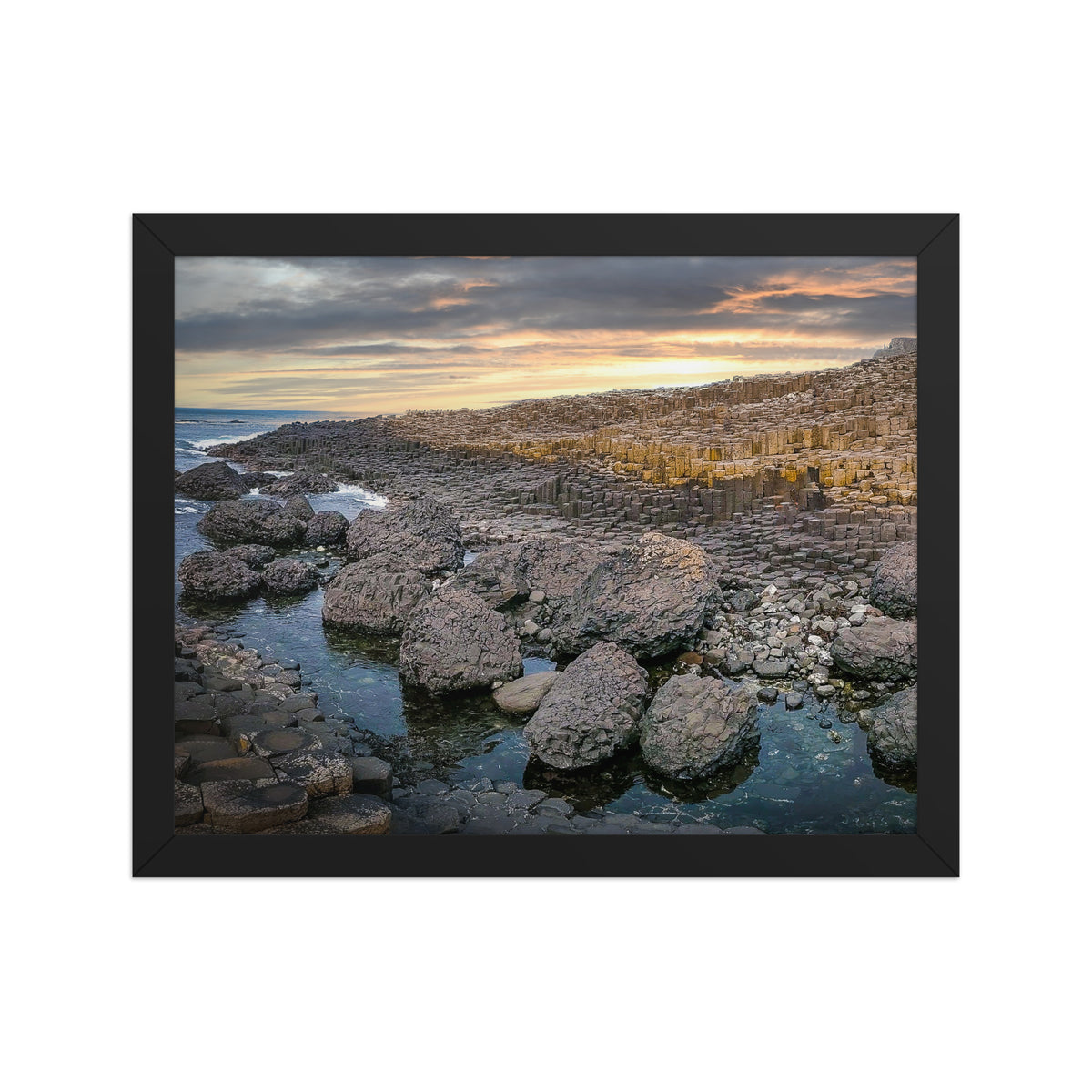 Giant's Causeway, Northern Ireland framed luster print by iain, front view of a black framed photograph of basalt columns and dark boulders along the ocean under a golden sunset sky
