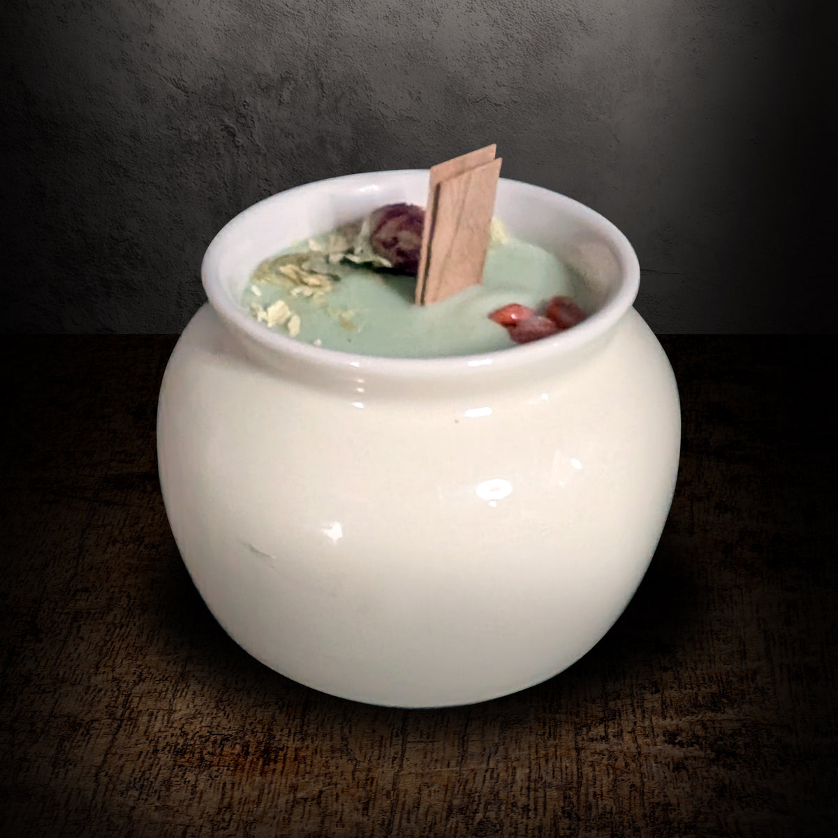 Cream porcelain pot candle on a dark background, pale green soy wax topped with dried red globe amaranth and white botanicals around a flat wooden wick