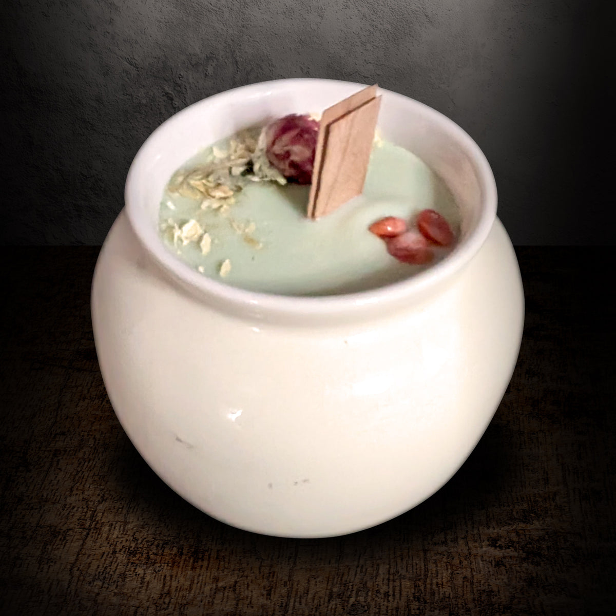 Cream porcelain pot candle seen from a high angle on a dark background, showing the pale green wax surface set with dried flowers and red agate