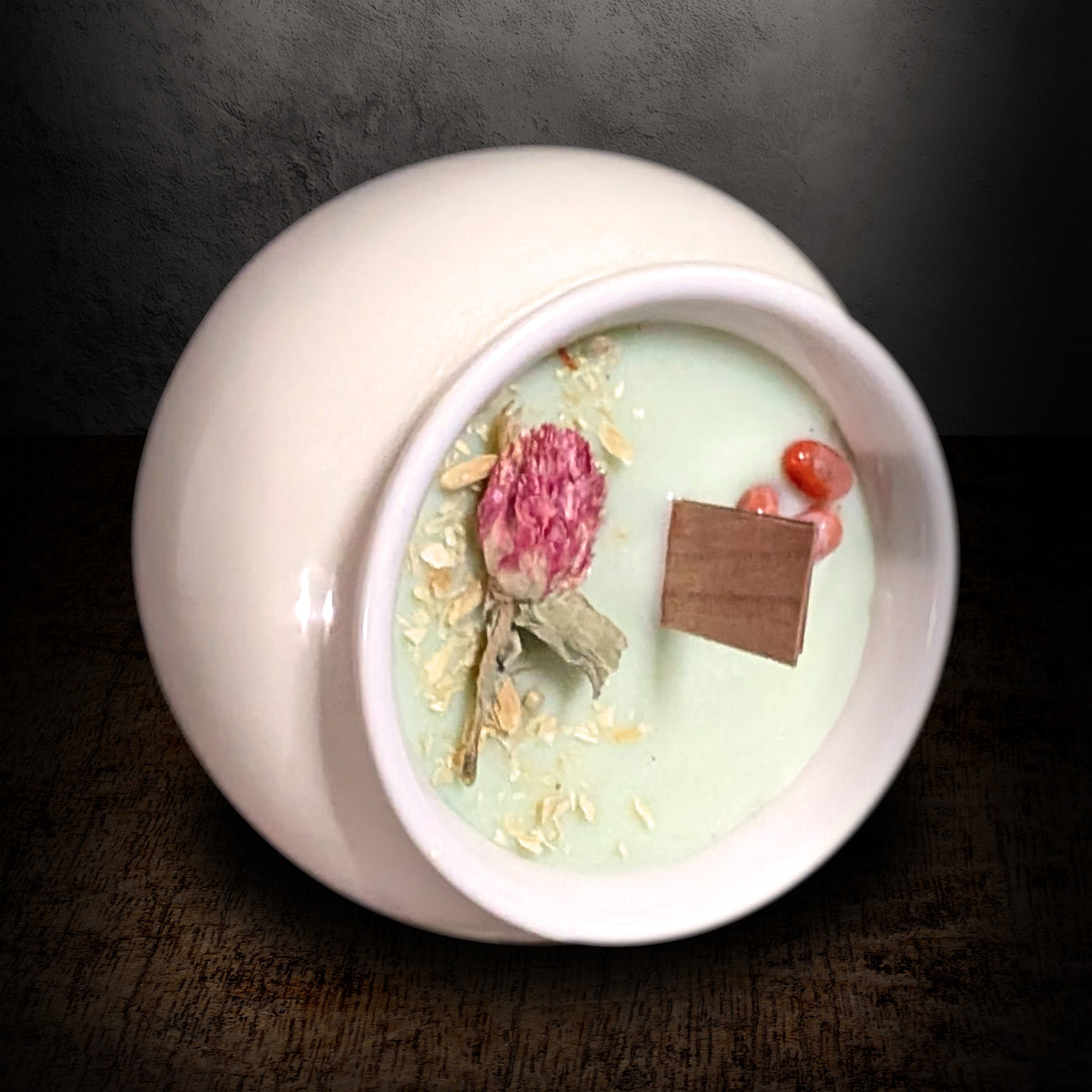 Cream porcelain pot candle tipped forward on a dark background, showing the full wax surface with dried red globe amaranth, white botanicals and red agate
