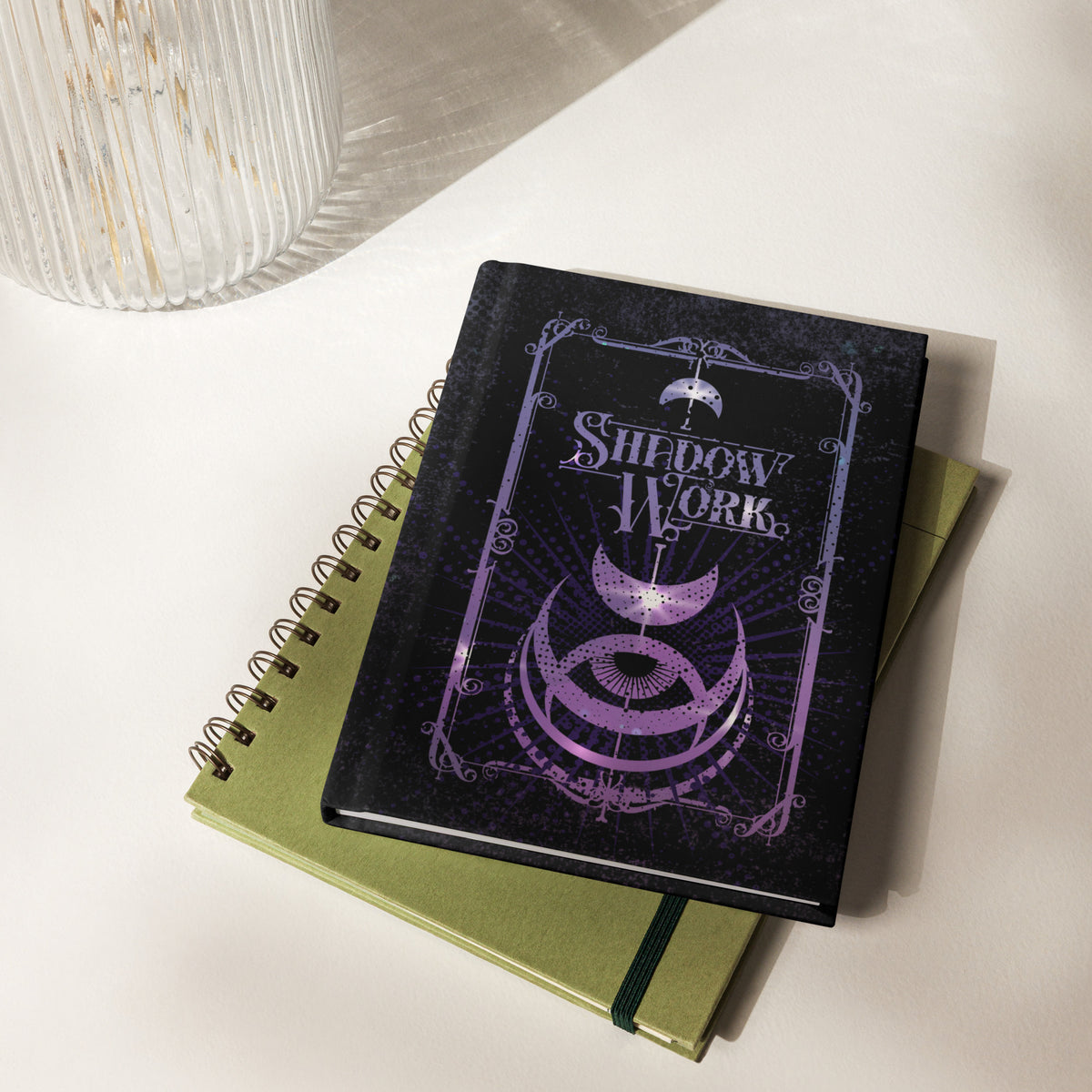 Shadow Work journal stacked on top of an olive green notebook on a white desk beside a fluted glass vase