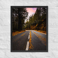 Mountain Road framed luster print by iain, black framed photograph of a winding road through a dense pine forest under a pink and violet sky, hanging on a white brick wall