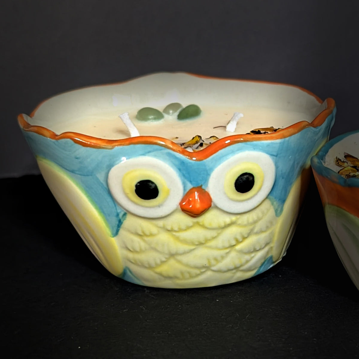 Close up of the larger owl candle on a dark background, blue brow and orange beak over a quilted pale green breast, wax surface set with green aventurine beside the wick
