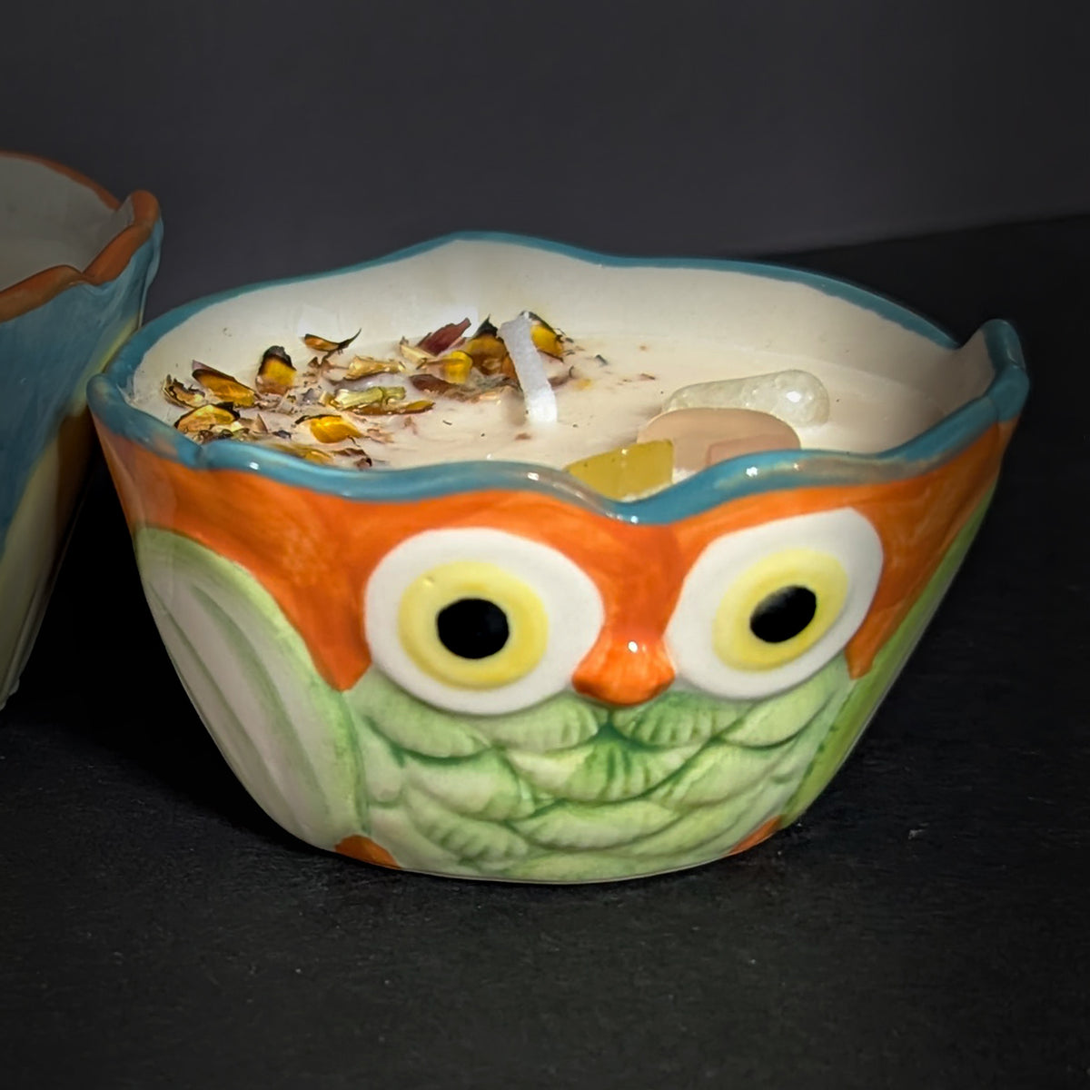 Close up of the smaller owl candle on a dark background, orange brow and blue eye band over a quilted pale green breast, wax surface scattered with dried chrysanthemum petals and stones