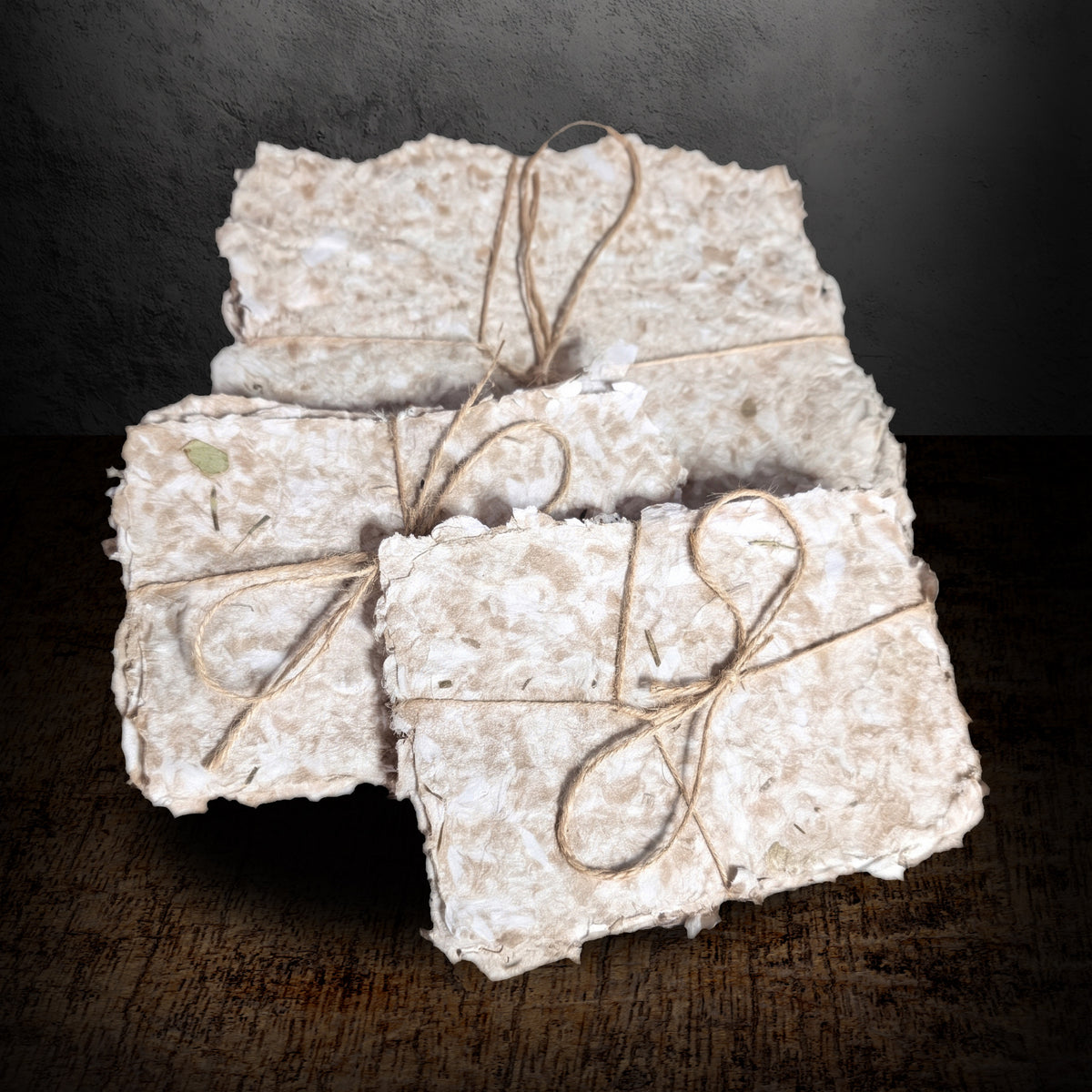 Bundles of handmade paper tied with twine, shown close up from the front on a dark background, rough deckle edges and herb flecks visible in the pulp