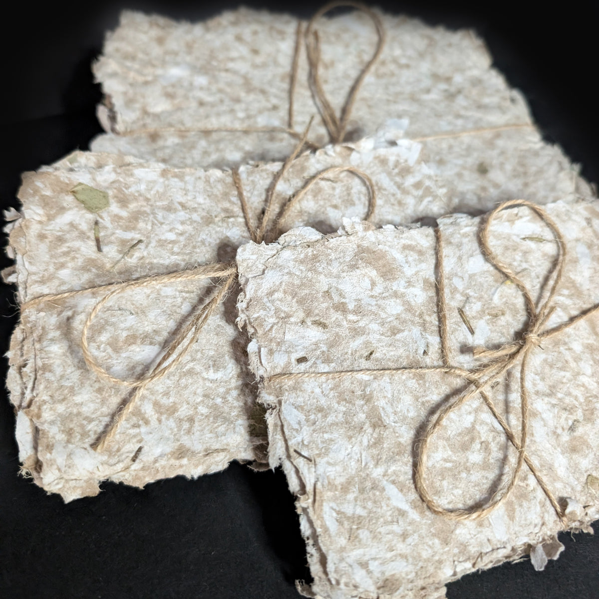 Close detail of the handmade paper surface on a dark background, showing the thick pressed pulp fibers and pieces of dried herb worked into the sheet