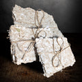 Three bundles of handmade paper tied with twine on a dark background, one standing upright behind the others, cream pulp with deckle edges and flecks of dried herbs