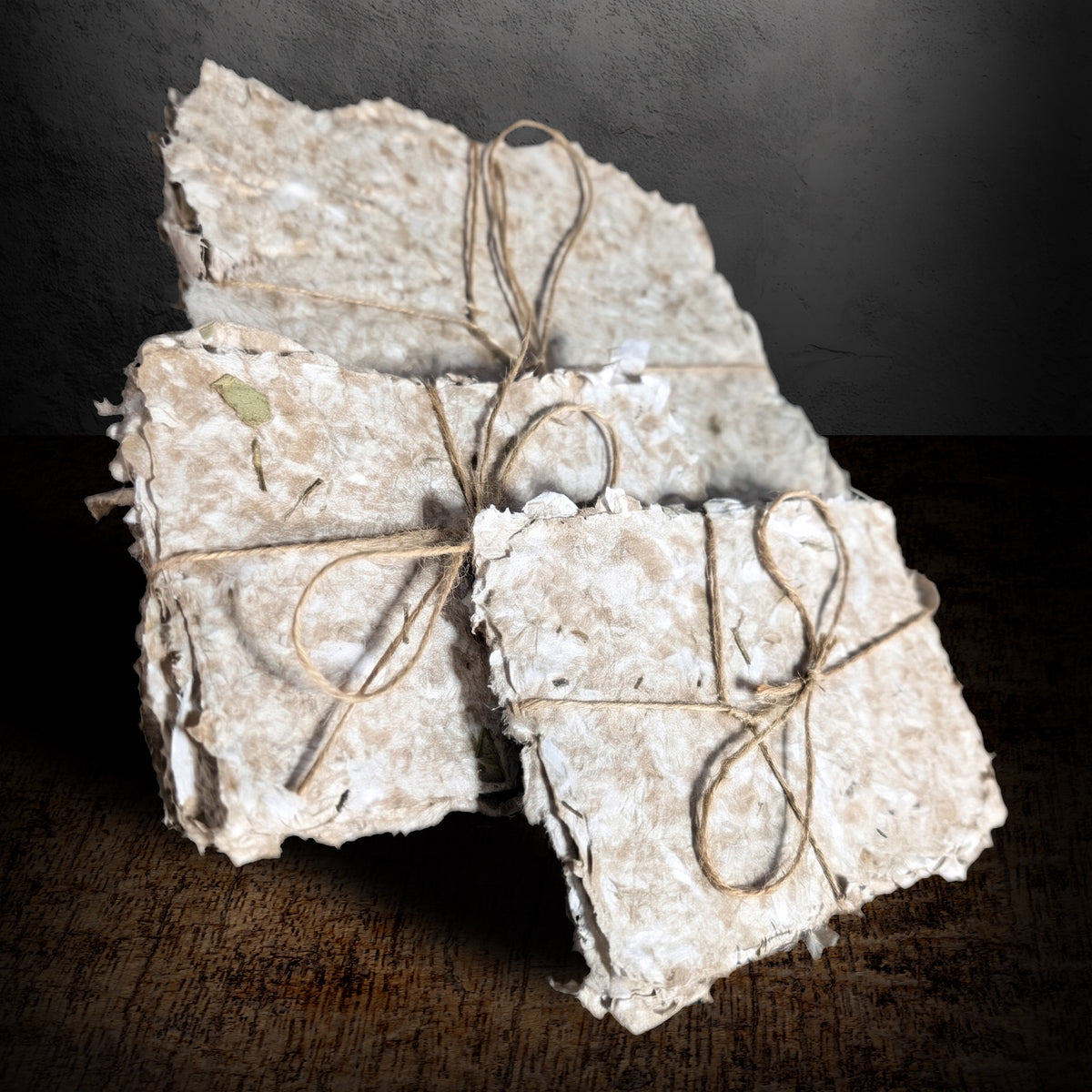 Three bundles of handmade paper tied with twine on a dark background, one standing upright behind the others, cream pulp with deckle edges and flecks of dried herbs