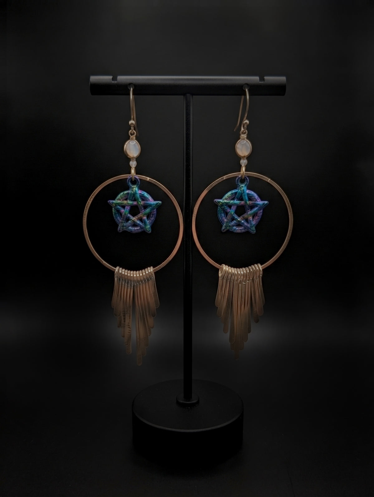 A pair of long bohemian-style dangle earrings displayed on a black stand against a dark background. Each earring has a small white bead, a bronze-toned hoop, a blue and purple iridescent pentagram charm, and a long bronze-toned fringe tassel.