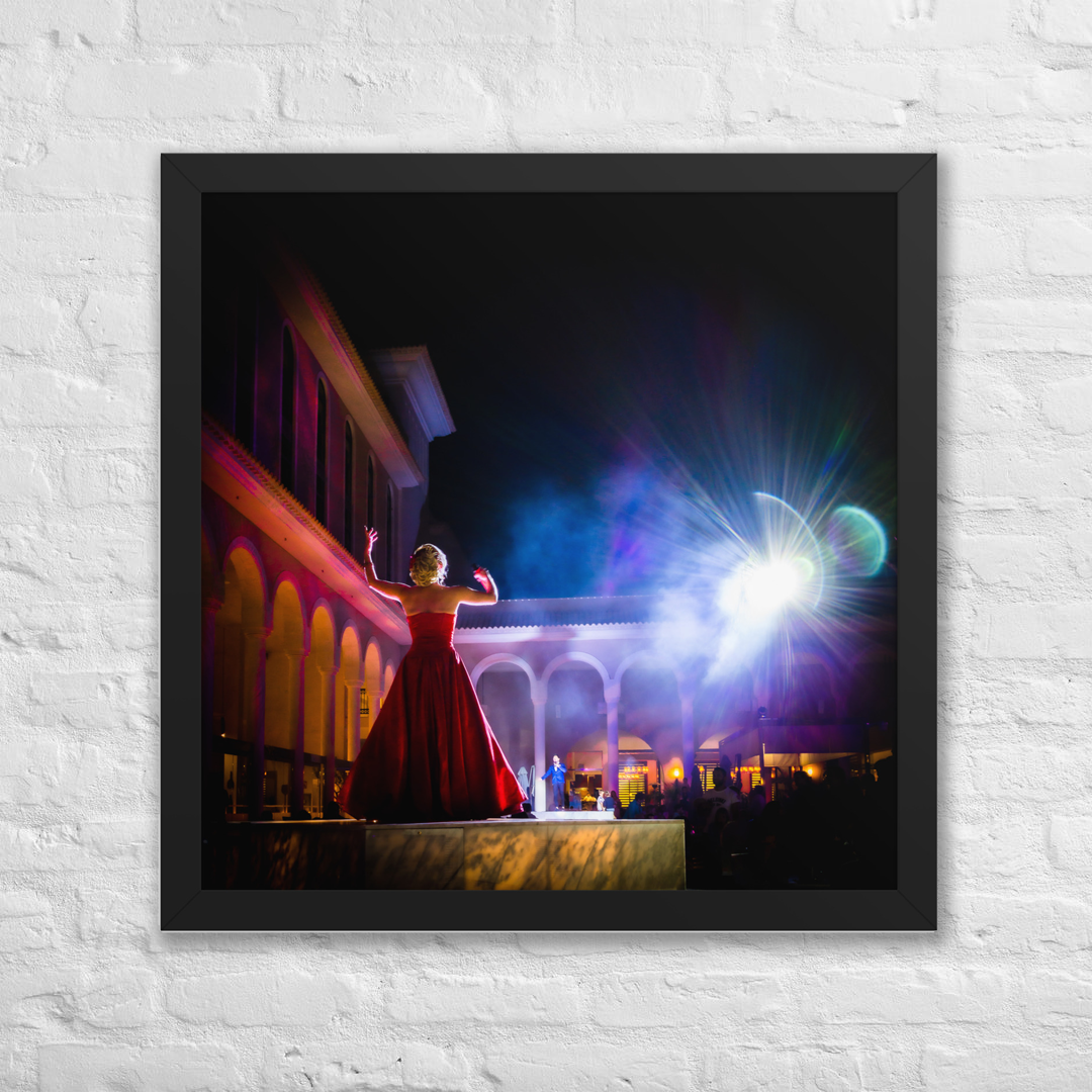 Perform for the Gods framed luster print by iain, large square black framed photograph of a performer in a red gown raising her arms toward the night sky, with glowing arches, smoke, and a burst of stage light, hanging on a white brick wall