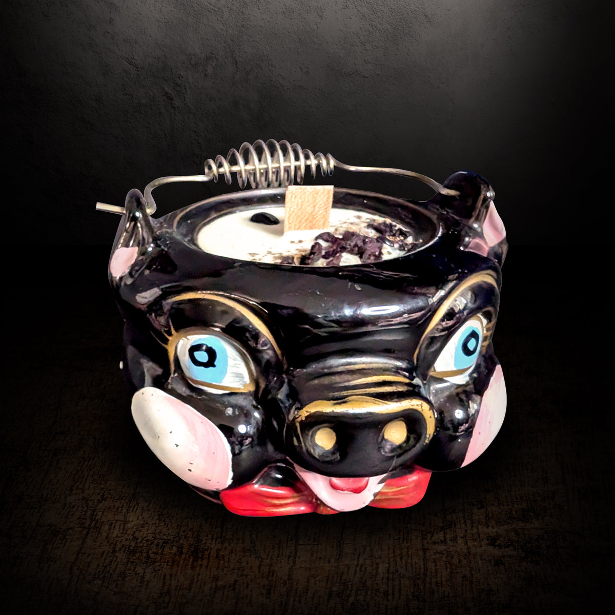 Black ceramic pig candle with painted blue eyes, pink snout and gold trim, seen from the front on a dark background, cream soy wax topped with obsidian chips and dried petals around a wooden wick