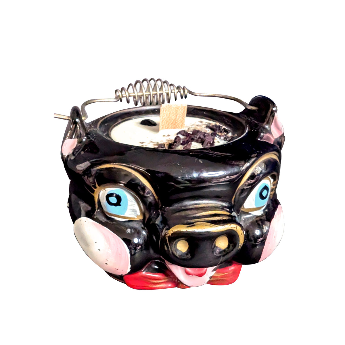 Black ceramic pig candle with painted blue eyes, pink snout and gold trim, seen from the front on a white background, cream soy wax topped with obsidian chips and dried petals around a wooden wick