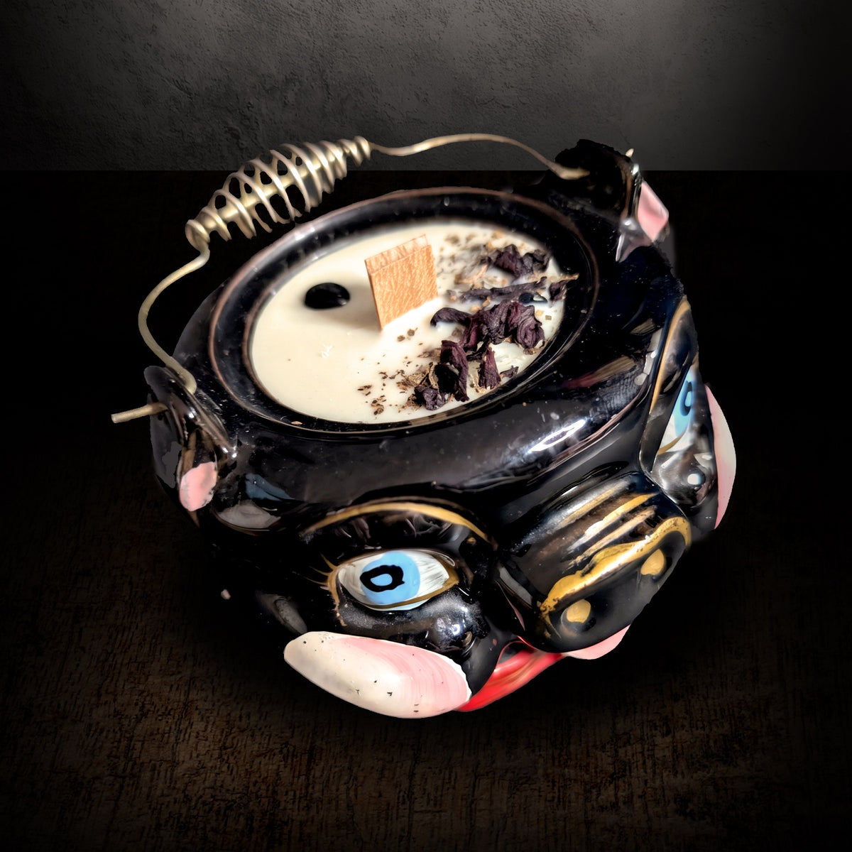Black ceramic pig candle seen from a high angle on a dark background, showing the wax surface set with obsidian chips and dried petals and the wire bail handle across the top