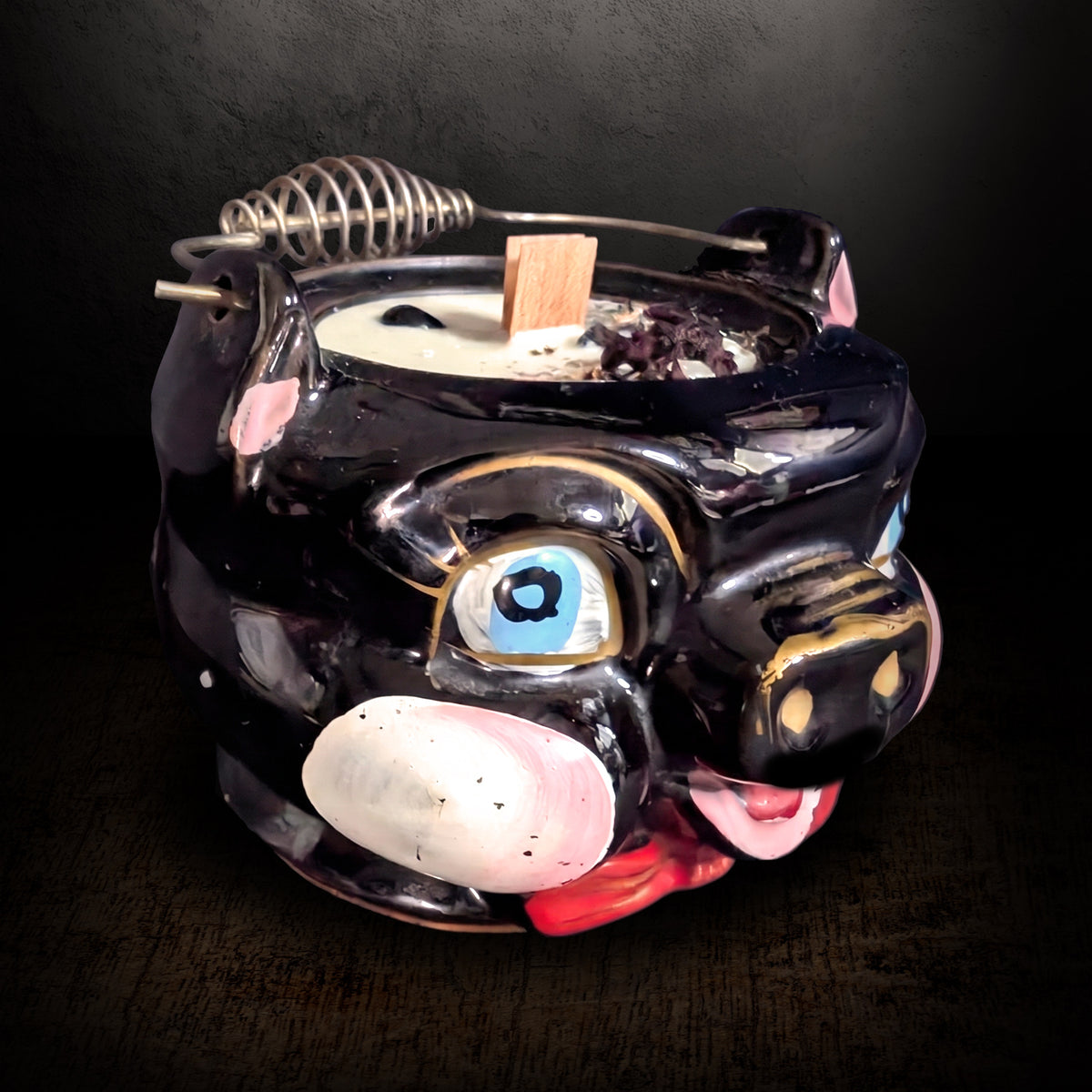 Black ceramic pig candle turned to the side on a dark background, showing the hand painted eye, pink snout and the wire bail handle