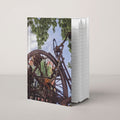 Rusty Bicycle hardcover journal by iain, journal standing upright on a soft neutral background, with a photograph of a rusted bicycle overgrown with roots and moss under green leaves and blue sky on the front cover and the page edges showing on the side