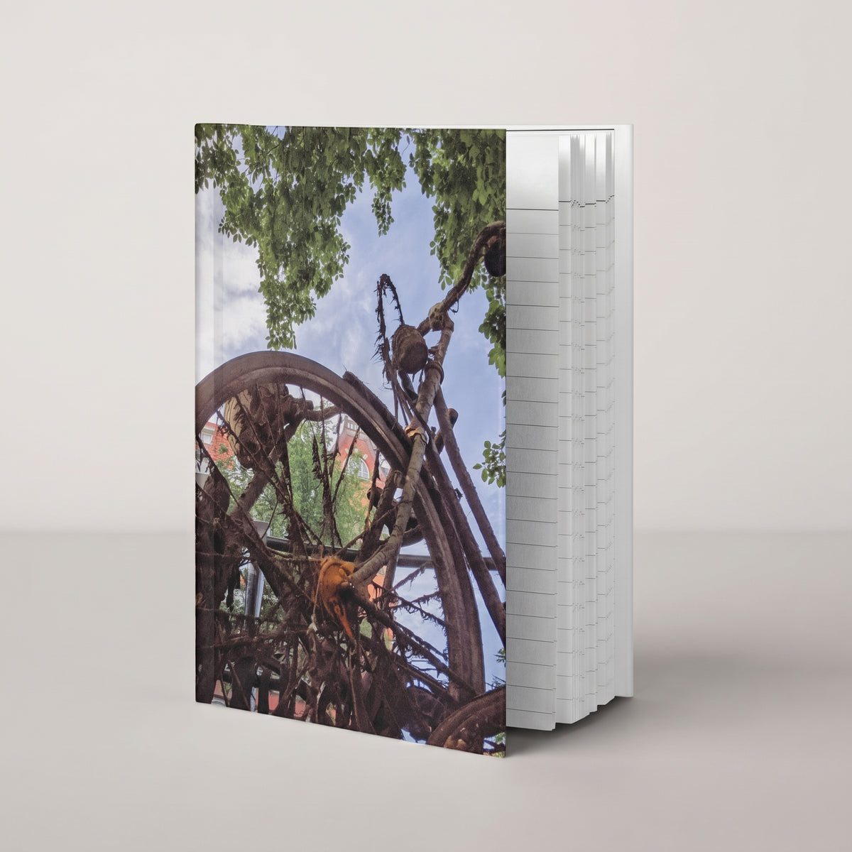 Rusty Bicycle hardcover journal by iain, journal standing upright on a soft neutral background, with a photograph of a rusted bicycle overgrown with roots and moss under green leaves and blue sky on the front cover and the page edges showing on the side