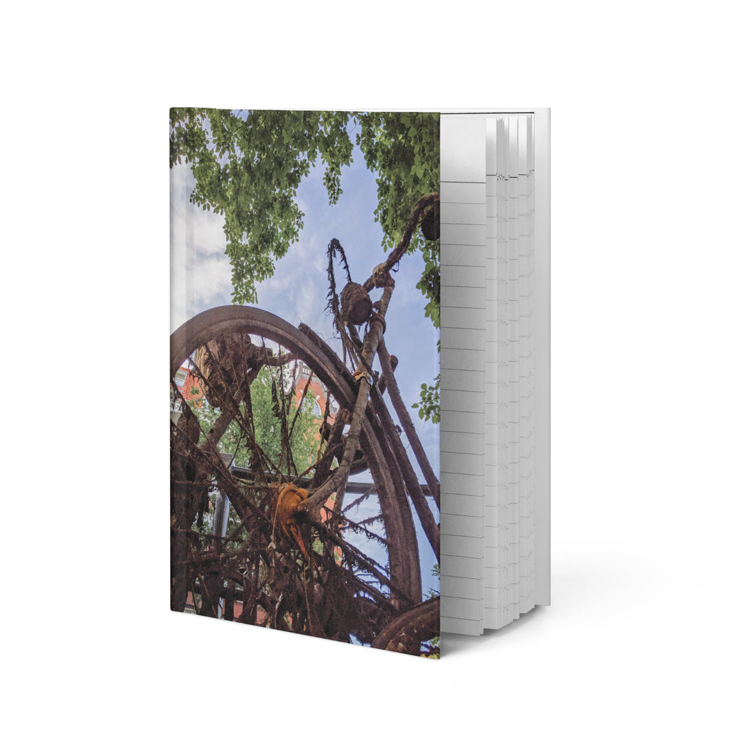 Rusty Bicycle hardcover journal by iain, journal standing upright on a white  background, with a photograph of a rusted bicycle overgrown with roots and moss under green leaves and blue sky on the front cover and the page edges showing on the side
