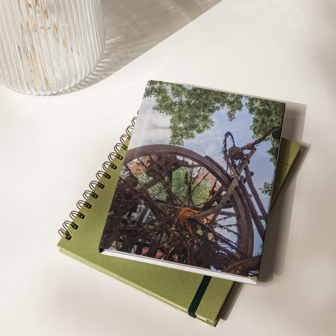 Rusty Bicycle hardcover journal by iain, journal stacked on a table, with a photograph of a rusted bicycle overgrown with roots and moss under green leaves and blue sky on the front cover and the page edges showing on the side