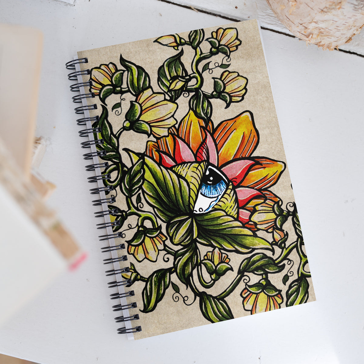 Watchful Nature spiral notebook lying flat, showing the full cover art of flowers, vines, and the blue eye at the center
