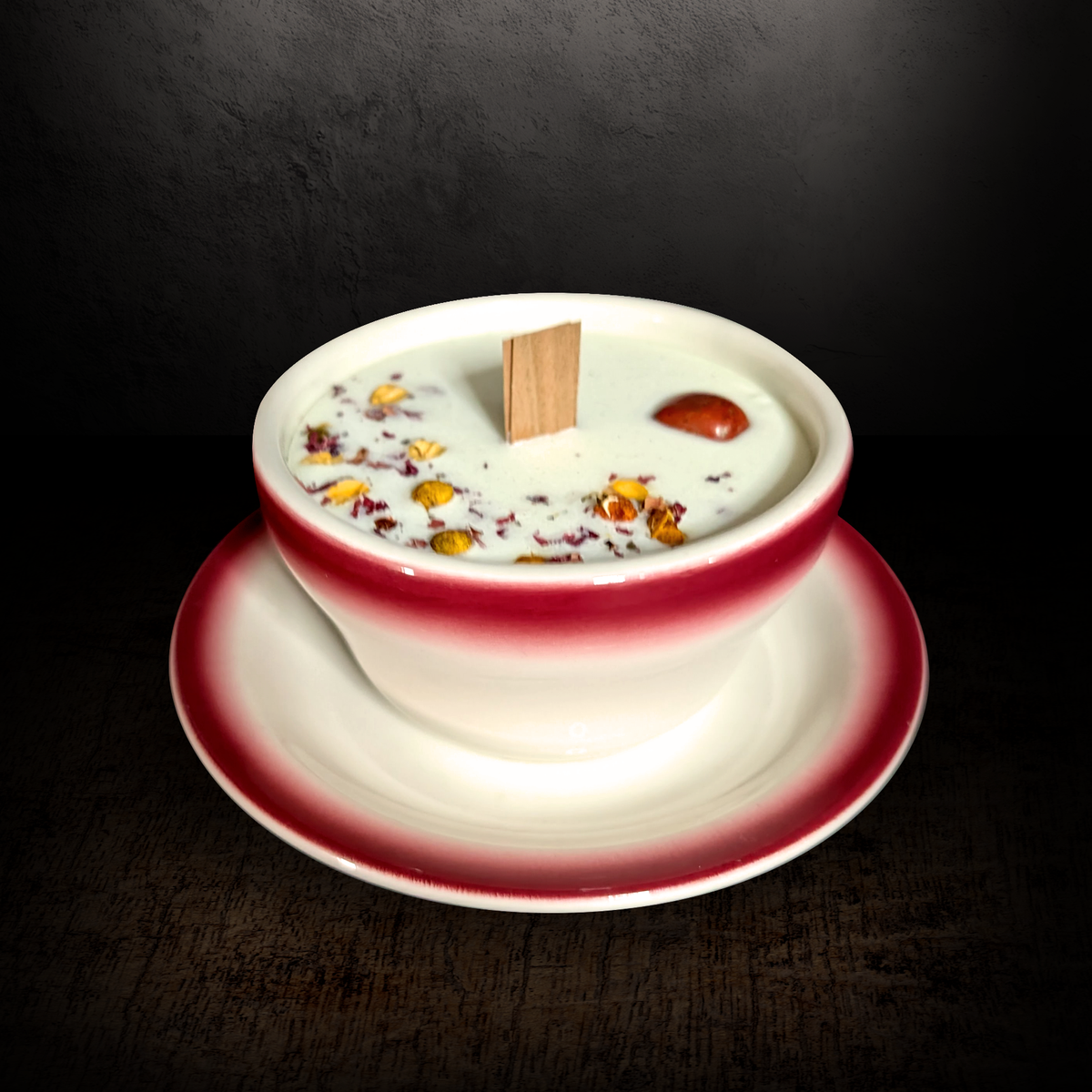Red and white teacup candle with saucer, front view on a dark background, white soy wax topped with dried botanicals and a flat wooden wick