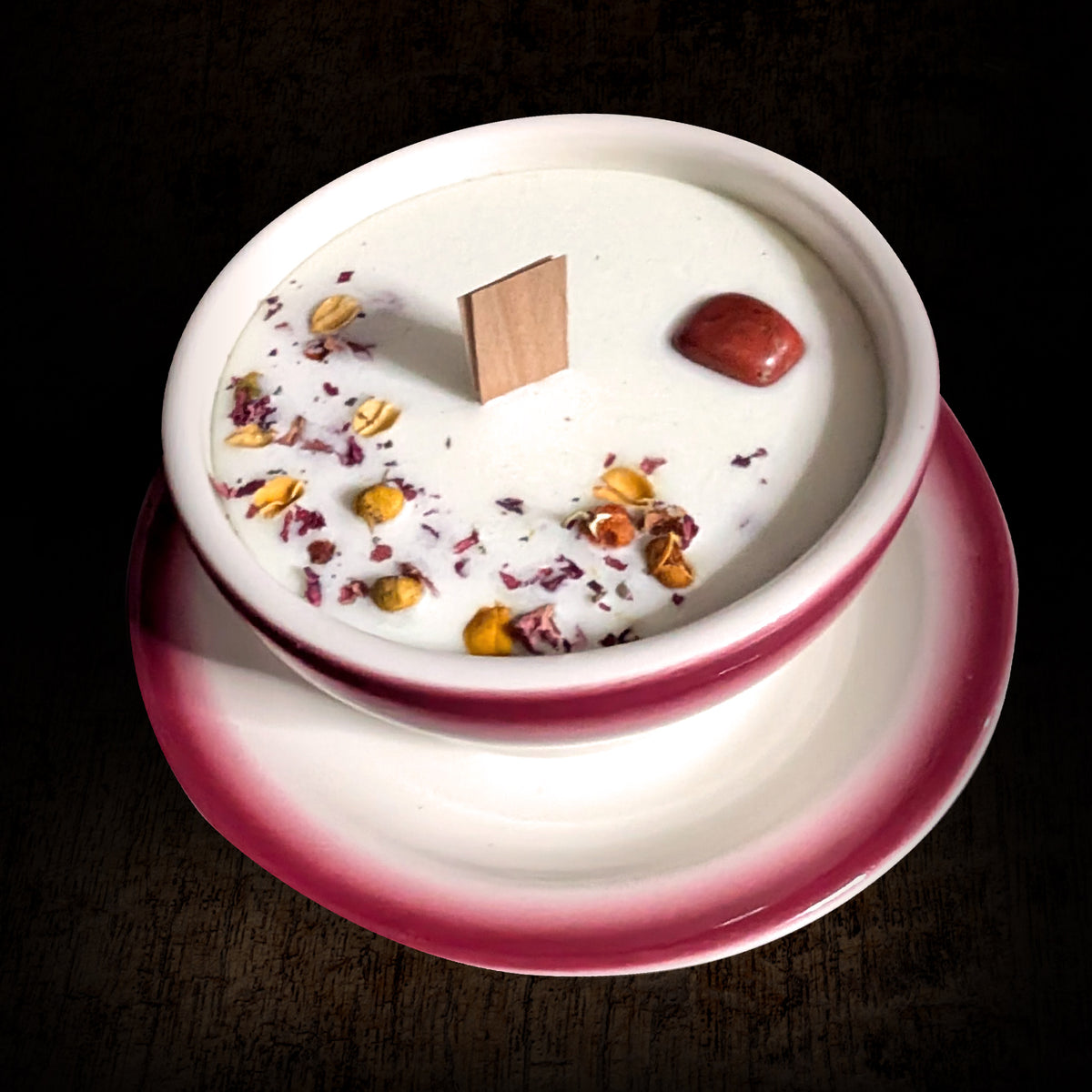 Red and white teacup candle seen from above on a dark background, dried flower petals and botanicals pressed into the wax surface
