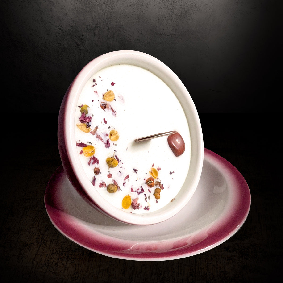 Red and white teacup candle tipped forward on its saucer on a dark background, showing the wax surface and wooden wick