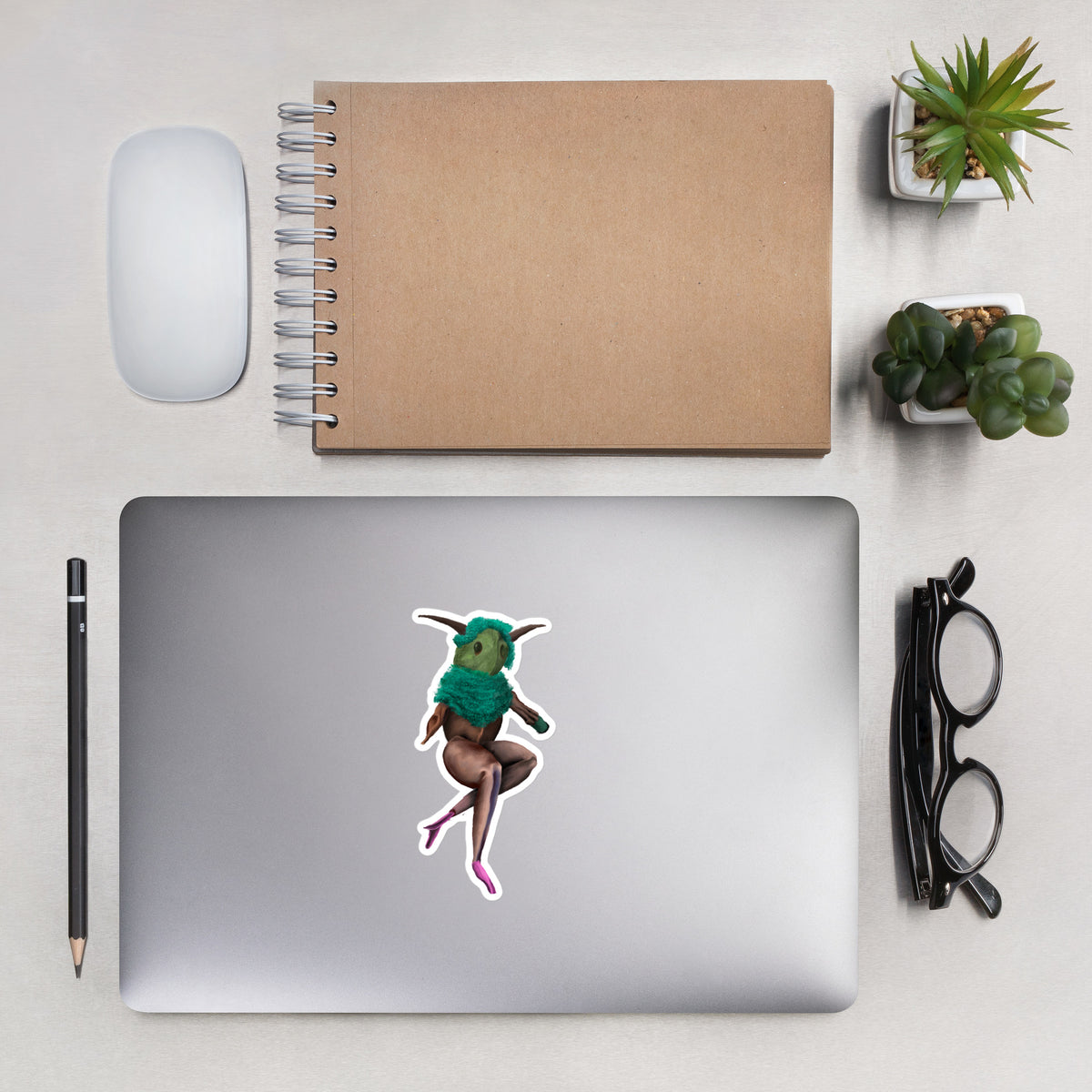 Vinyl sticker applied to the lid of a silver laptop on a desk setup with a notebook, pen, and glasses.