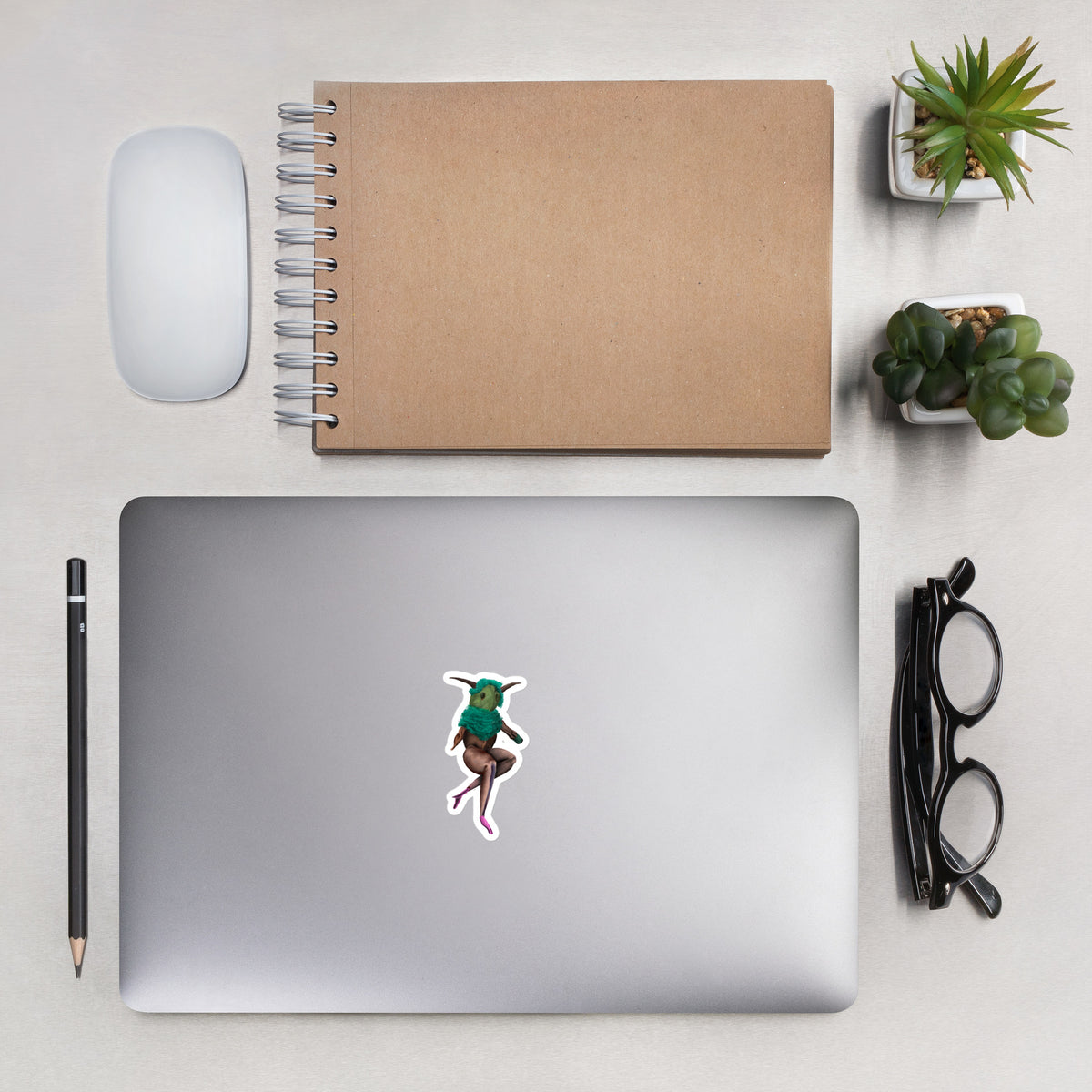 Overhead lifestyle view of the sandpile sprite sticker placed on a laptop next to a notebook and desk accessories to show scale.