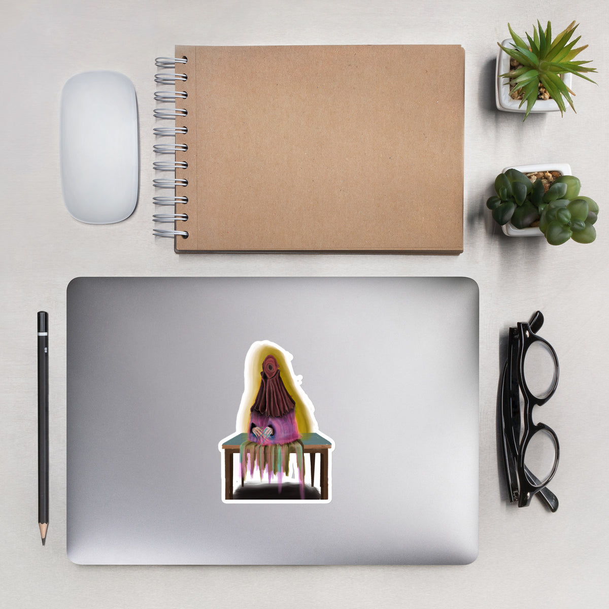 Matte vinyl sticker applied to the lid of a silver laptop on a desk setup with a notebook, pen, and glasses.