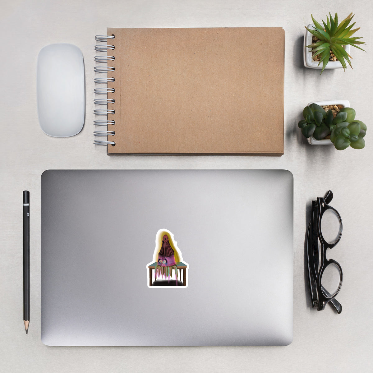 Overhead lifestyle view of the seated sprite sticker placed on a laptop next to office accessories to demonstrate relative scale.