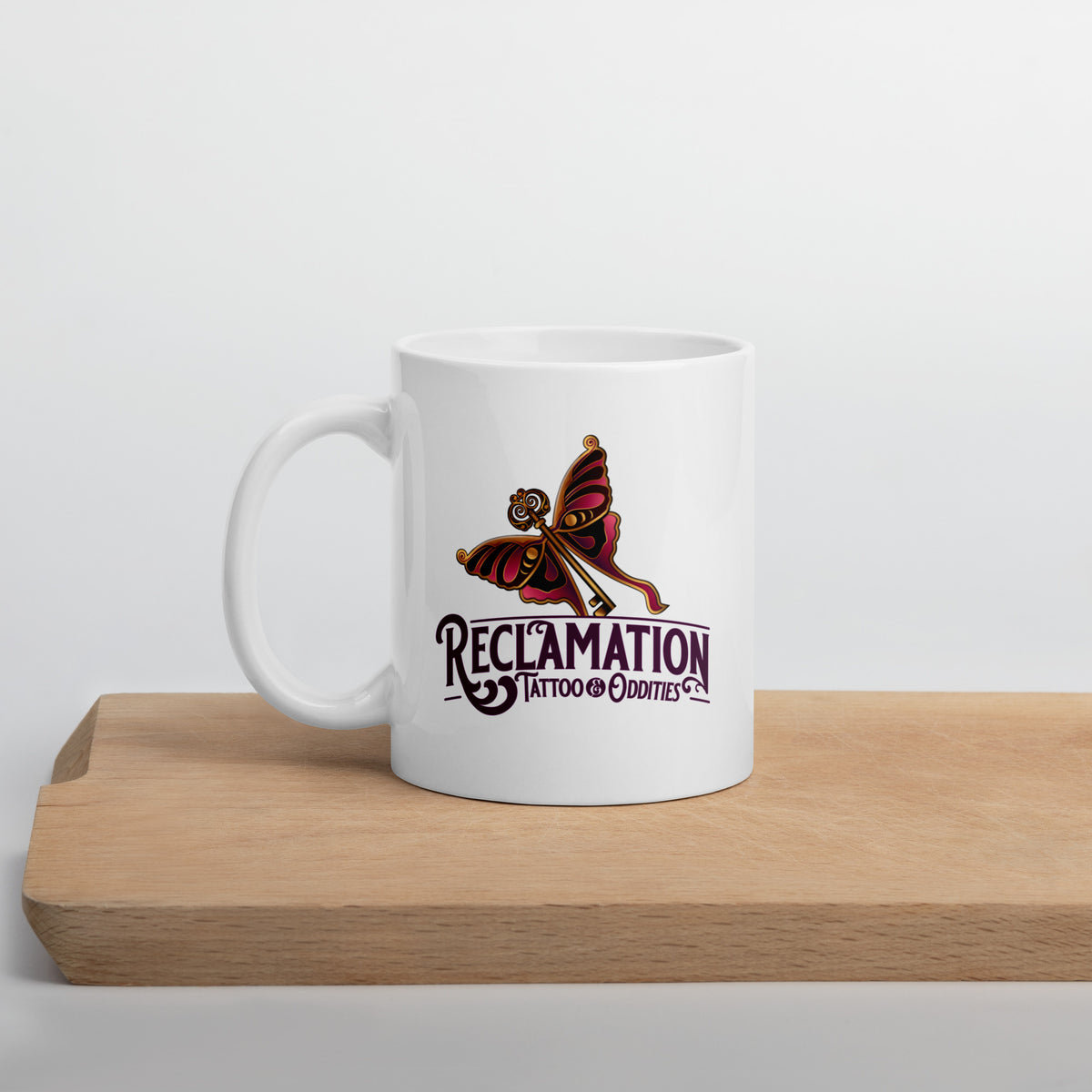 Reclamation & Midnight River mug in white resting on a wooden cutting board, the Reclamation Tattoo & Oddities logo facing the camera