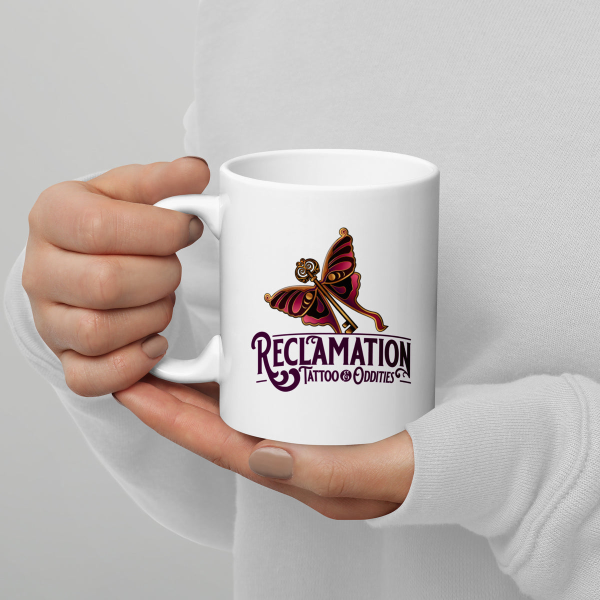 Reclamation & Midnight River mug in white held in a hand, handle on the left, the Reclamation Tattoo & Oddities butterfly and key logo facing the camera