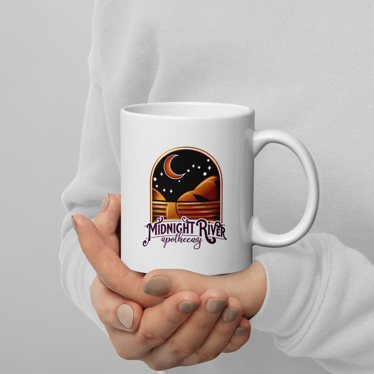 Reclamation & Midnight River mug in white held in a hand, handle on the right, the Midnight River Apothecary badge facing the camera