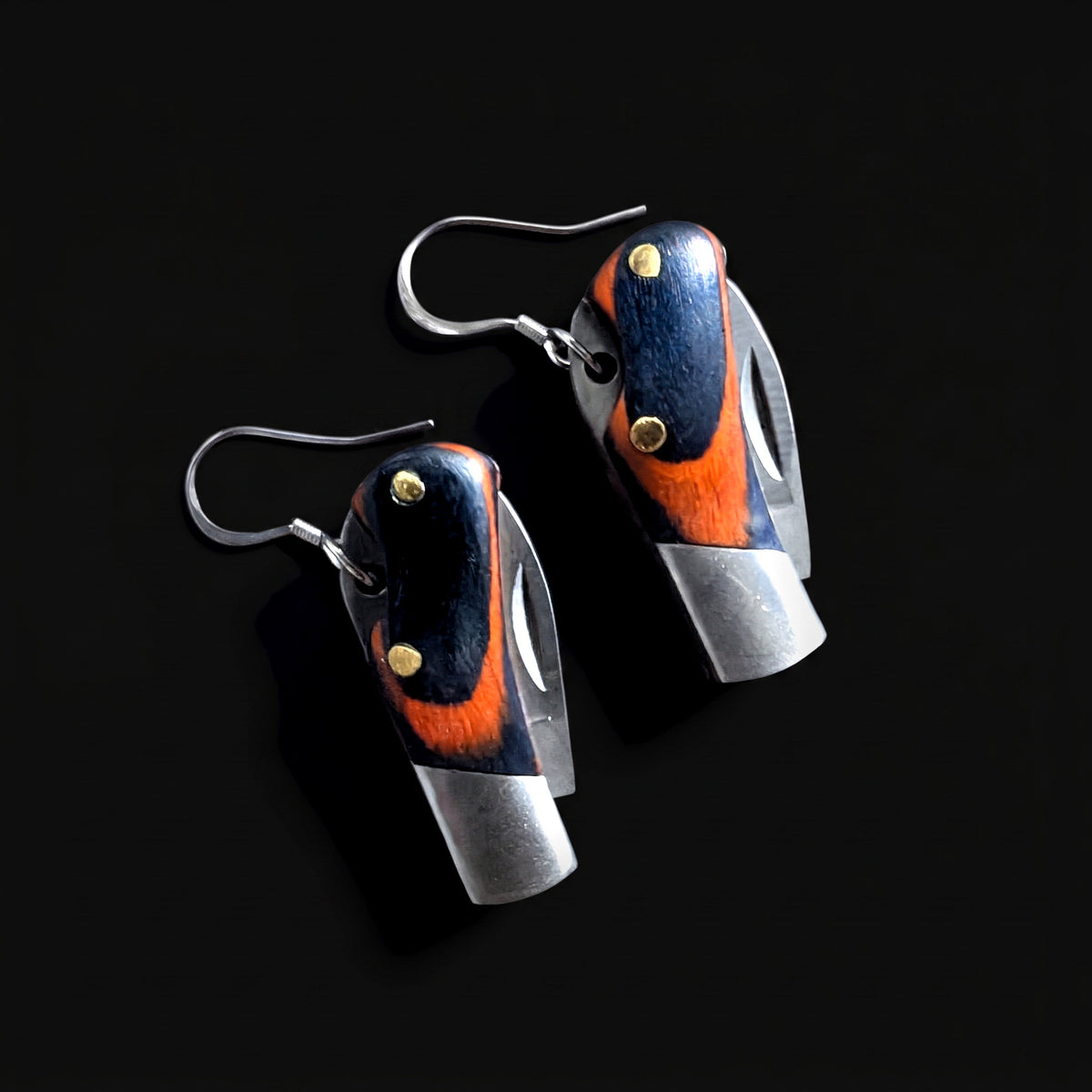 Pair of mini pocket knife earrings with layered orange and dark blue wood handles, brass pins and open steel blades, hanging from silver ear wires on a dark background